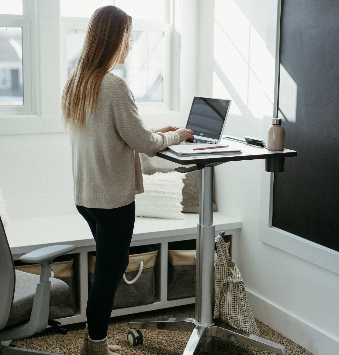 Young woman with long dirty-blonde hair, wearing black leggings, fuzzy socks and a comfy sweater is working on her laptop at home facing away from us, standing at Ergotron LearnFit Sit Stand Desk Mobile Cart. Light pours in from window.
